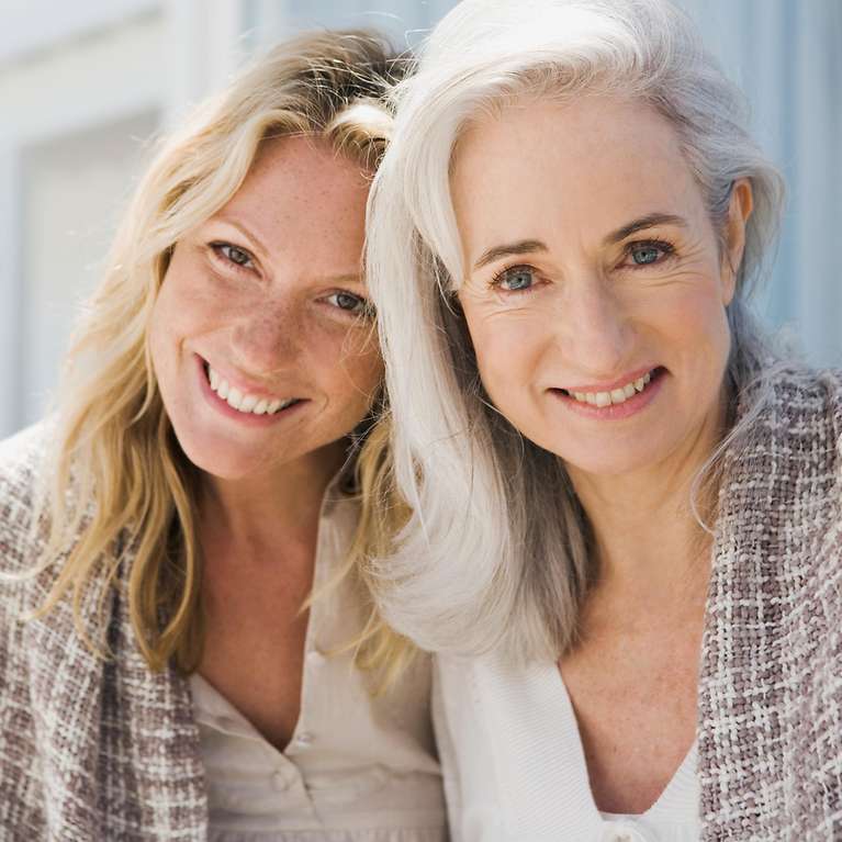 Zwei Frauen sitzen vor einem Haus und lächeln in die Kamera.