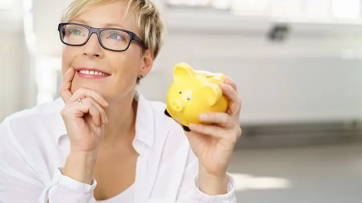 Eine Frau schaut lächelnd in die Ferne mit einem Sparschwein in der Hand.