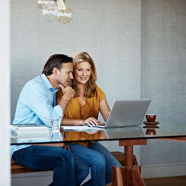 Eine Frau und ein Mann sitzen zusammen an einem Glastisch und schauen auf einen aufgeklappten Laptop.