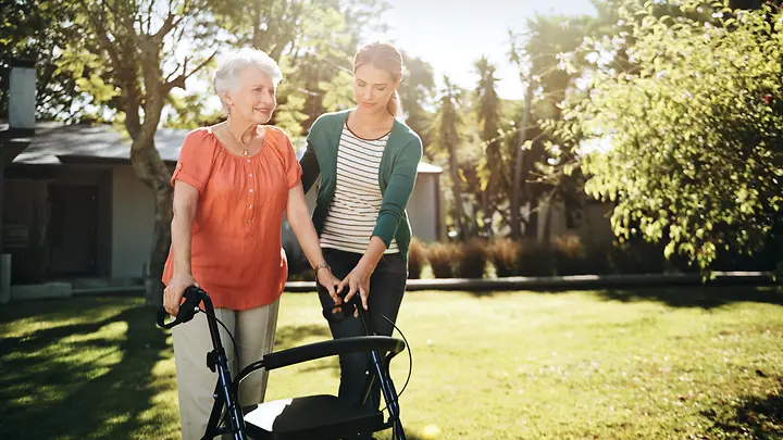 Man sieht eine Frau, die einer älteren Frau mit Gehhilfe unterstützt.