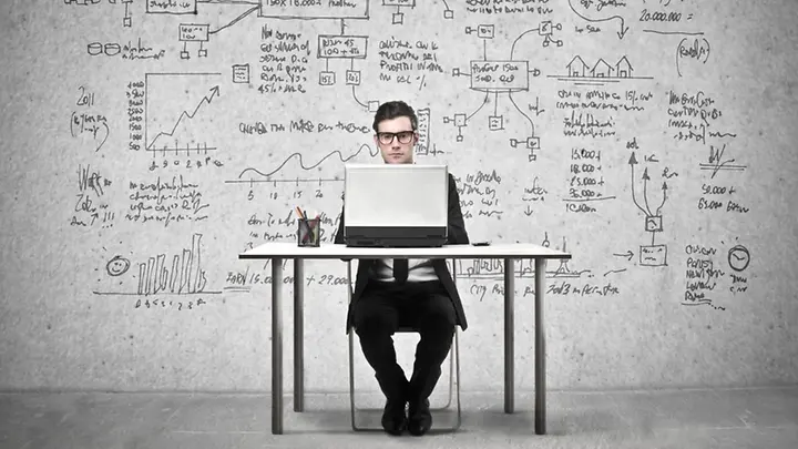 Ein Mann sitzt an einem Schreibtisch und schaut auf einen Laptop. Die Wand im Hintergrund ist vollgeschrieben.