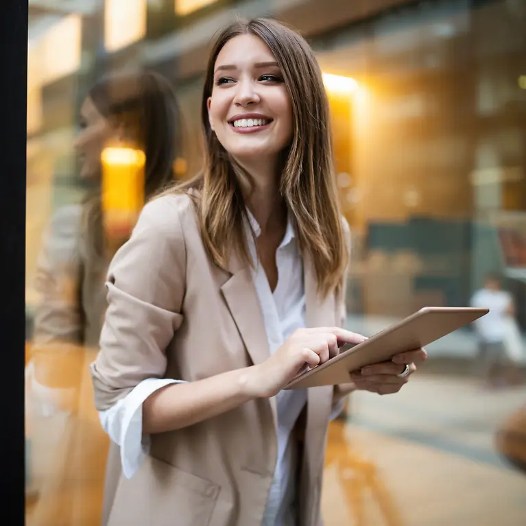 Eine Frau steht vor einem Fenster und hält ein Tablet in der Hand und lächelt.