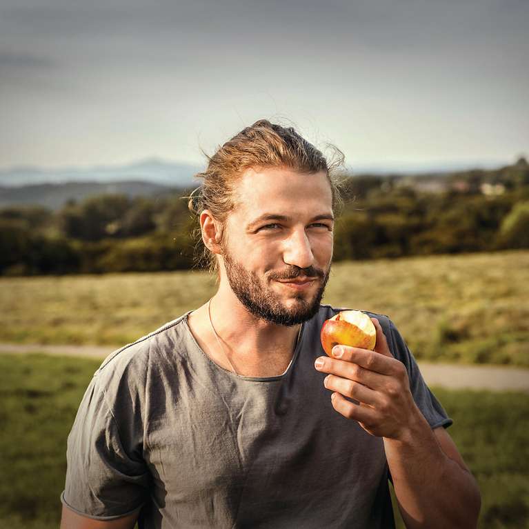 Ein Mann steht im Grünen, lächelt in die Ferne und hält einen Apfel in der Hand.