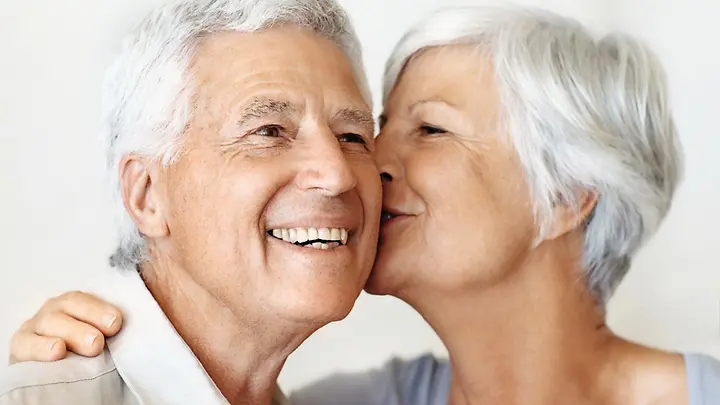 Man sieht ein ältere Dame, die ihrem Mann einen Kuss auf die Wange gibt.